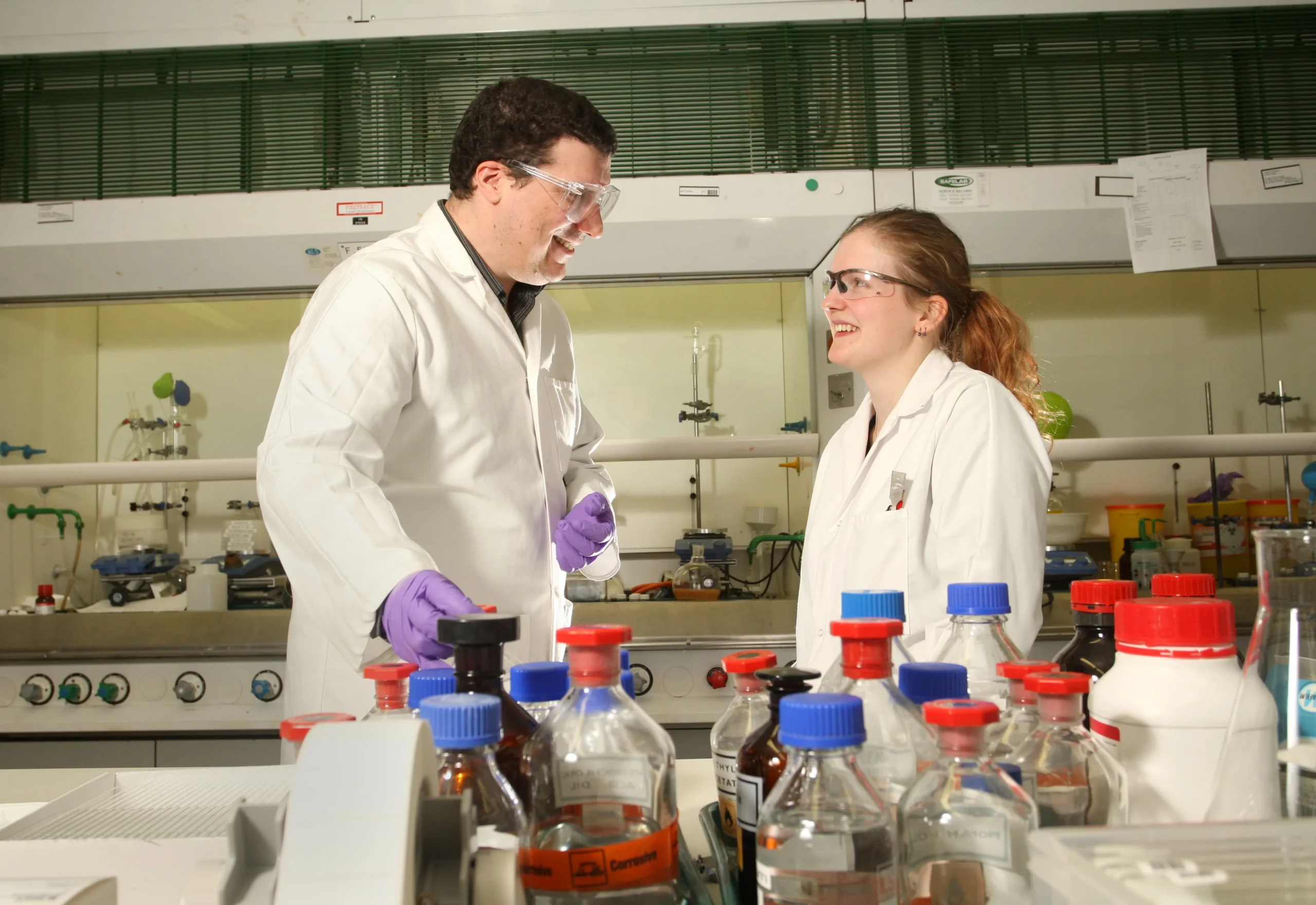 Two scientists in a laboratory discussing synthetic chemistry processes with glassware and reagent bottles on the bench, illustrating collaborative research and optimized reaction conditions.