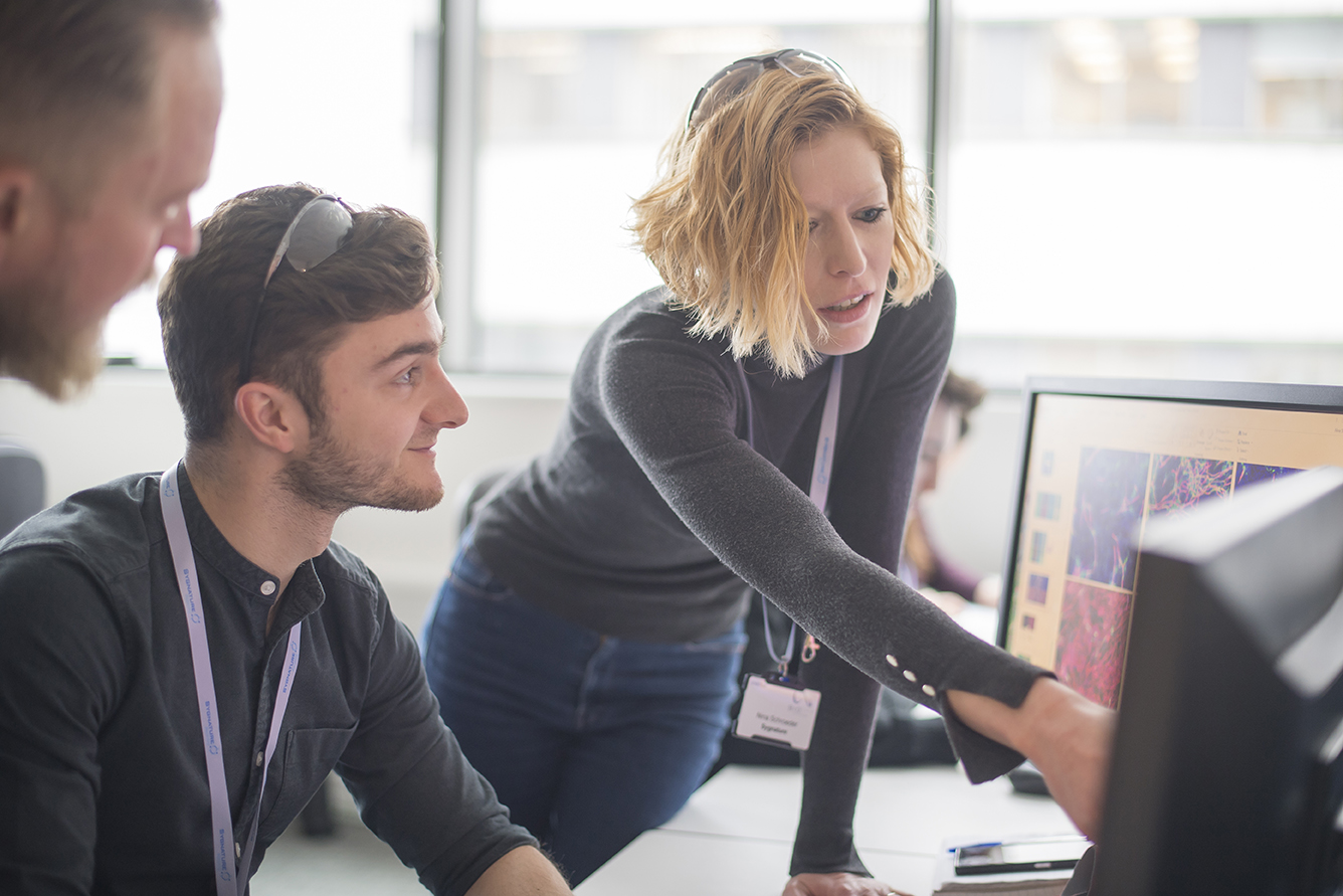 Environnement de bureau avec des membres d'équipe collaborant autour d'un écran d'ordinateur affichant un logiciel de gestion des flux de travail, représentant un flux de travail DMTA centralisé pour le suivi de la synthèse et l'analyse intégrée.