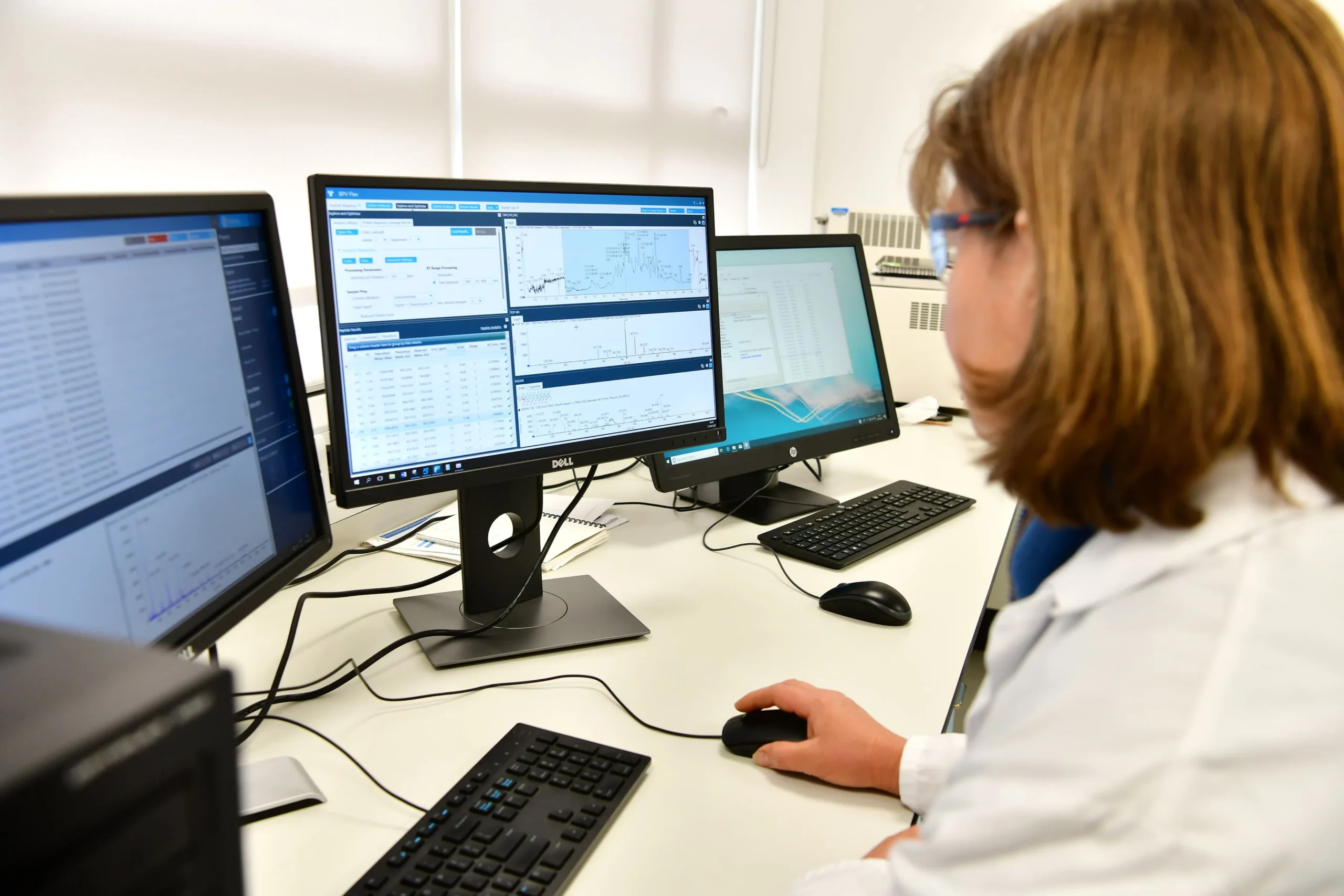 Laboratory workstation with mass spectrometry system and multiple monitors displaying data analysis for protein characterization