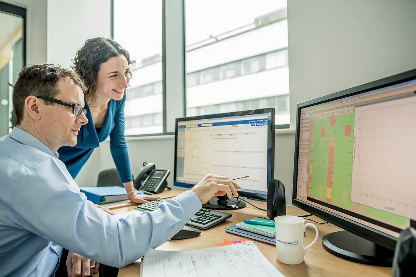 two Sygnature Discovery scientists reviewing complex data on dual computer monitors, supporting lead optimization and IND-enabling studies in drug discovery.