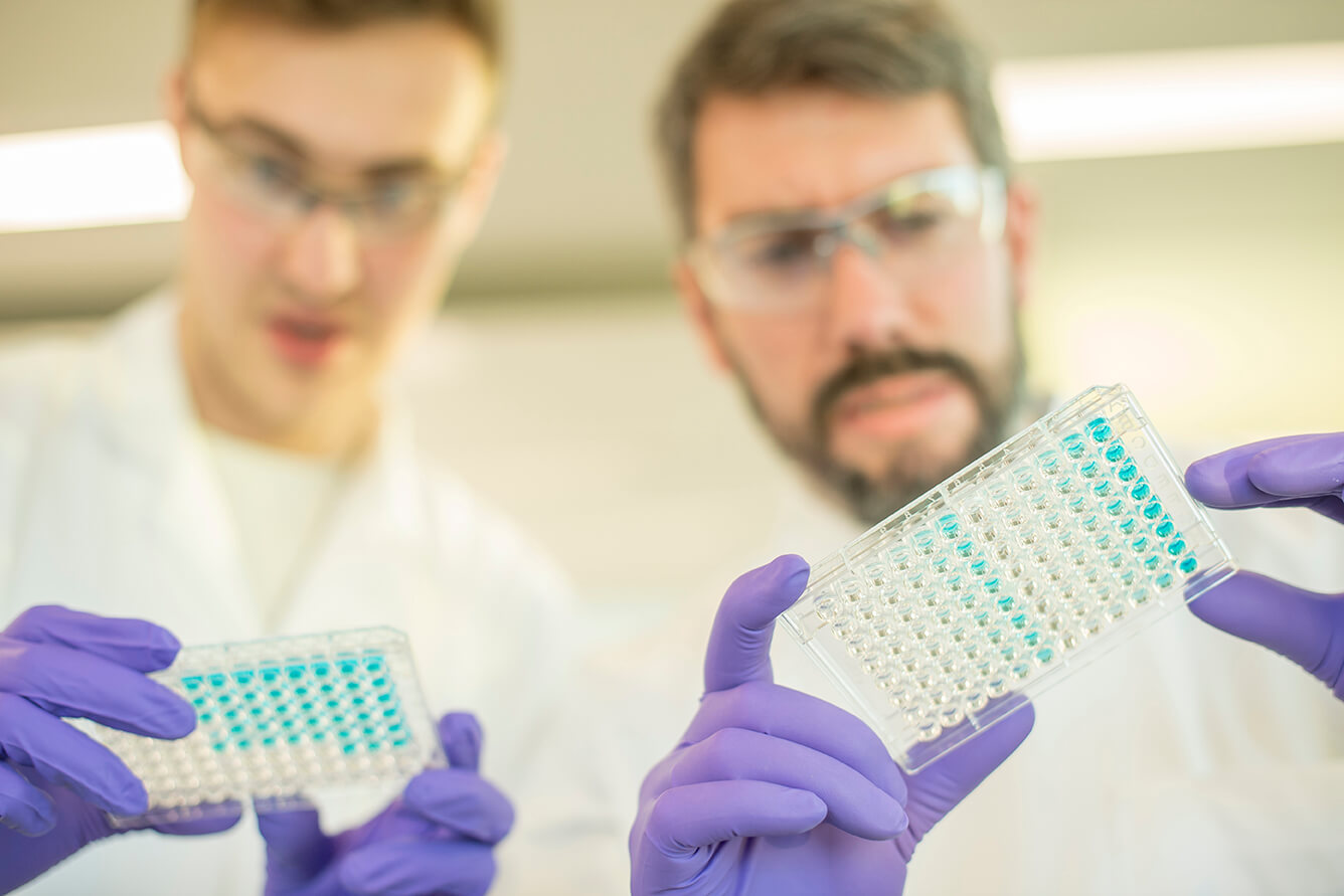 Close-up of gloved hands holding a microplate in a laboratory setting, representing metabolic disease research and drug discovery assays.