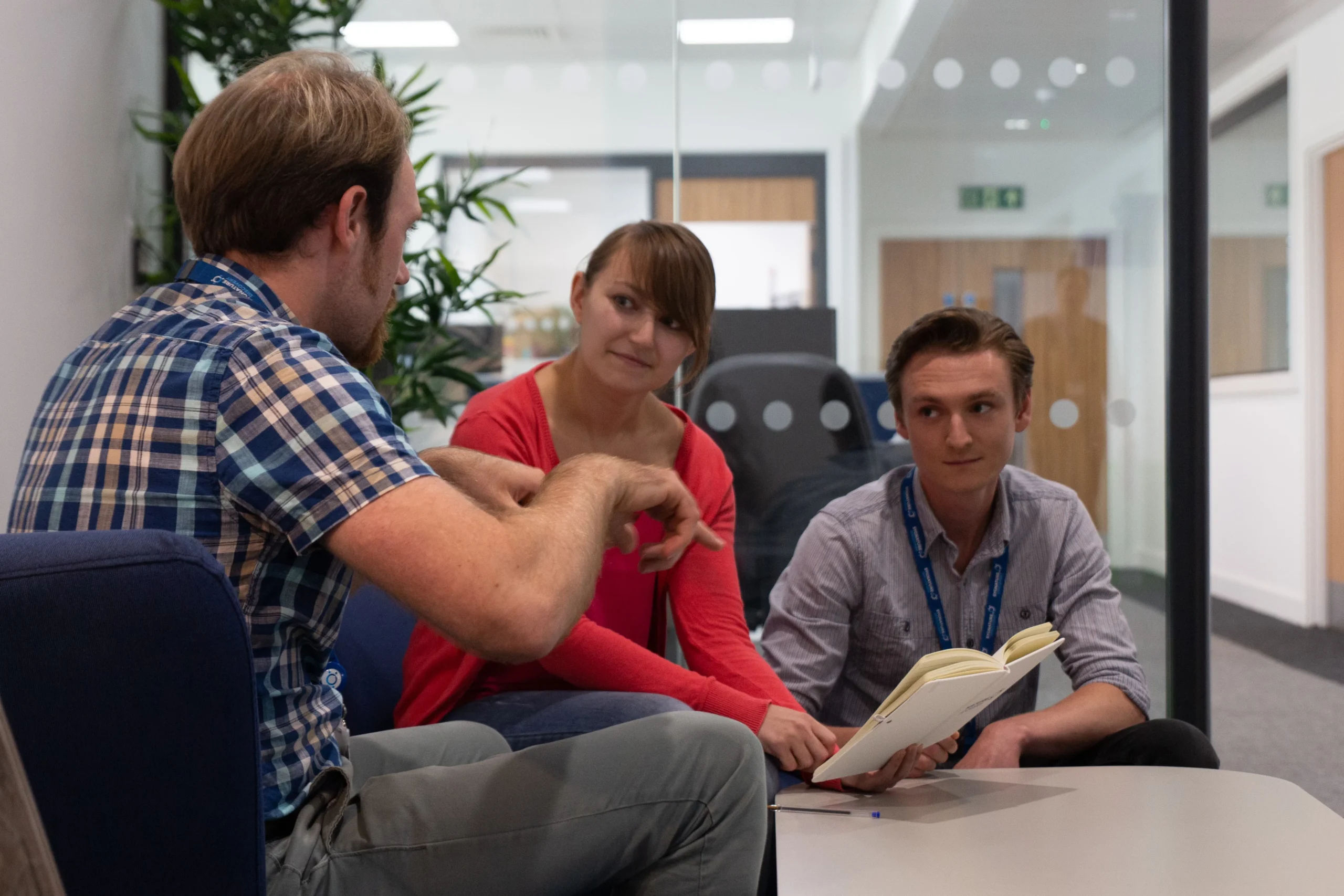 Three individuals seated in a modern office setting engaged in a discussion with an open notebook, representing tailored collaboration for ADME study design, PK/PD interpretation, and predictive modeling in drug discovery.